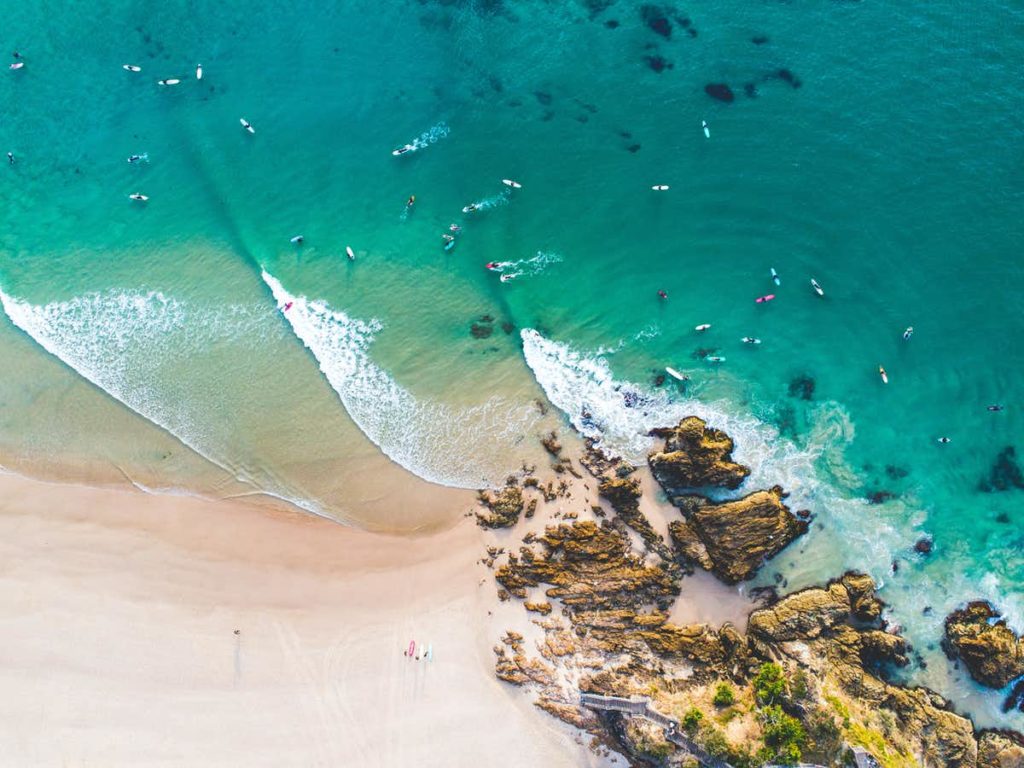 An aerial shot of the ocean, with clear blue water and a white sandy beach and colourful surfboards.