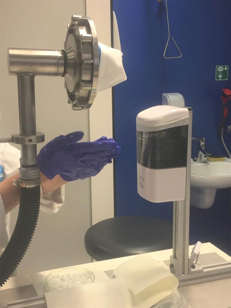 A lab replication of sanitising hands while wearing a face mask. Someone is rubbing sanitiser on gloved hands below a metal instrument that's holding a face mask. 