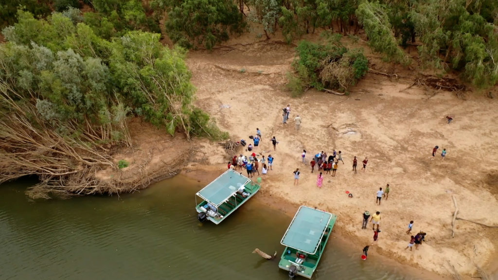 Indigenous seasonal calendars cover the Fitzroy Valley. River scene from above with two boats and groups of Indigenous people.