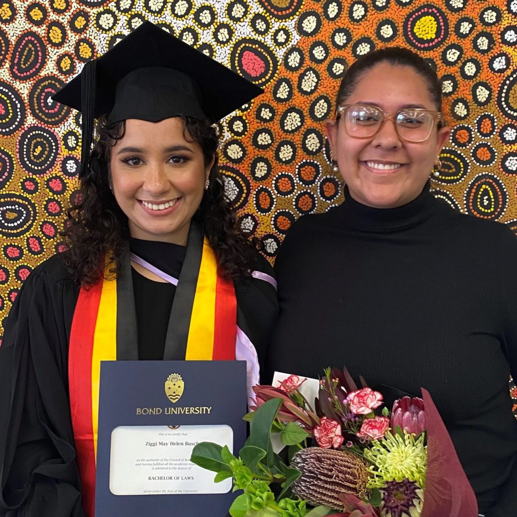 Maya Johnson and Ziggi Busch standing in front of an Indigenous motif artwork at Ziggi's graduation.