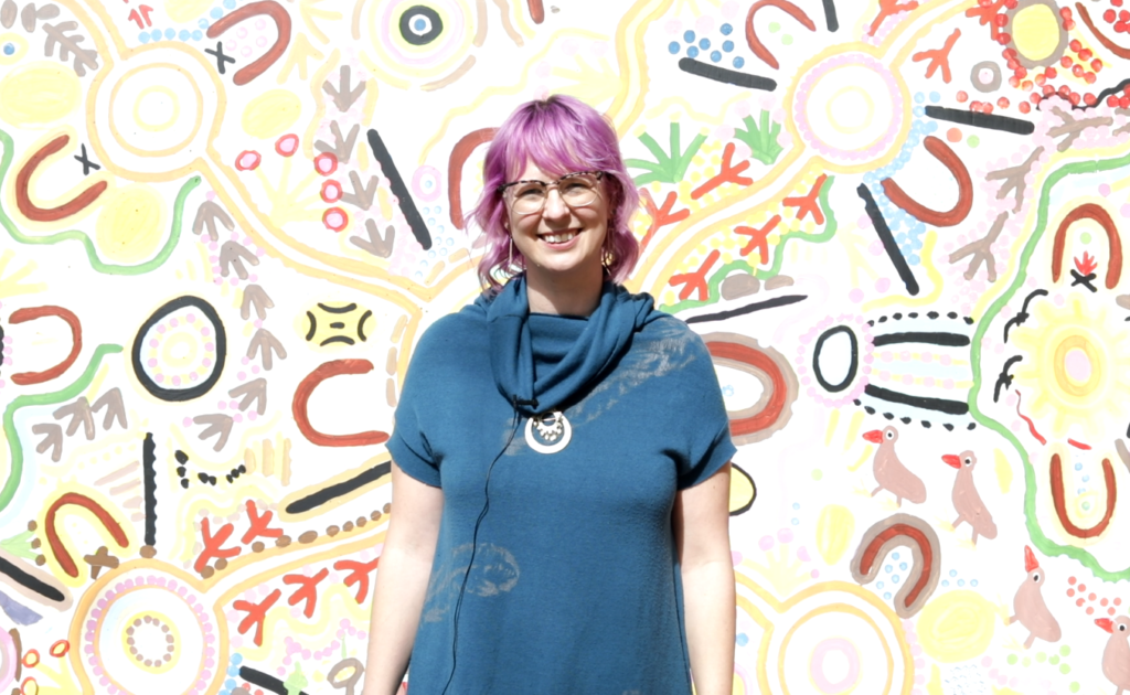 Nina Welti, wearing a blue jumper and has pink hair, is standing in front of a colourful, Indigenous painting wall.