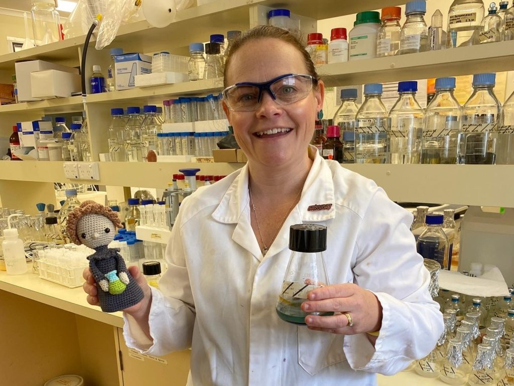 Waste innovation researcher Naomi Boxall standing in a lab holding a beaker and a crocheted doll