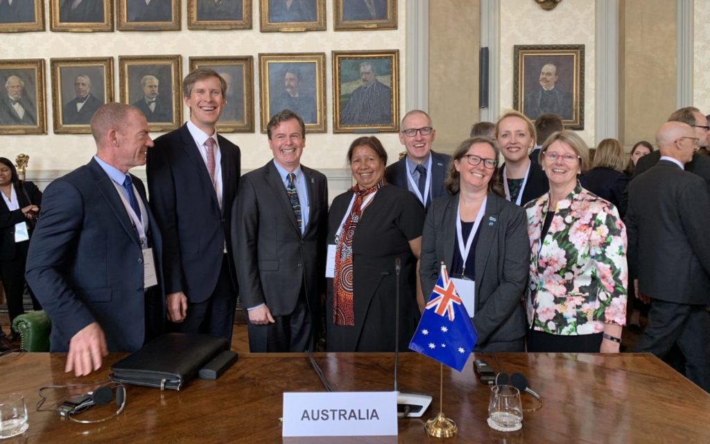 A group of men and women in formal attire pose behind a desk with a small Australian flag on it.