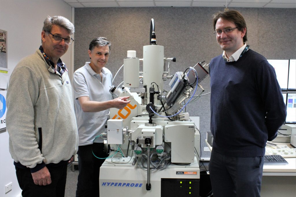 Three men standing in a lab next to an electron probe microanalyser (EPMA). 