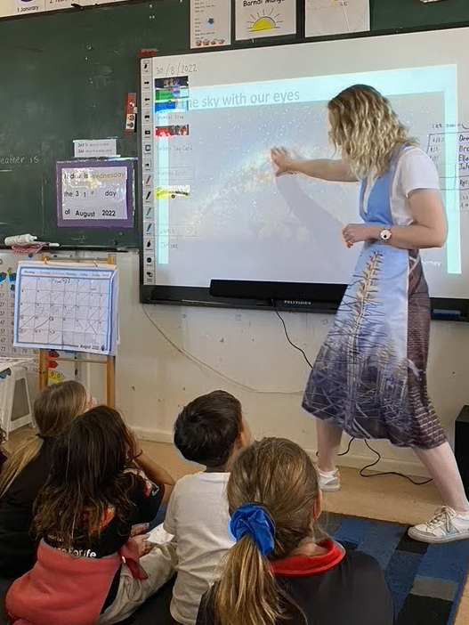 Laura standing in front of the class pointing at a screen that shows an image of the milky way. 