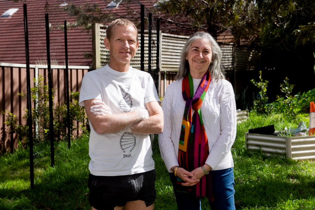 A man and a woman stand side by side in a garden and smile for the camera.