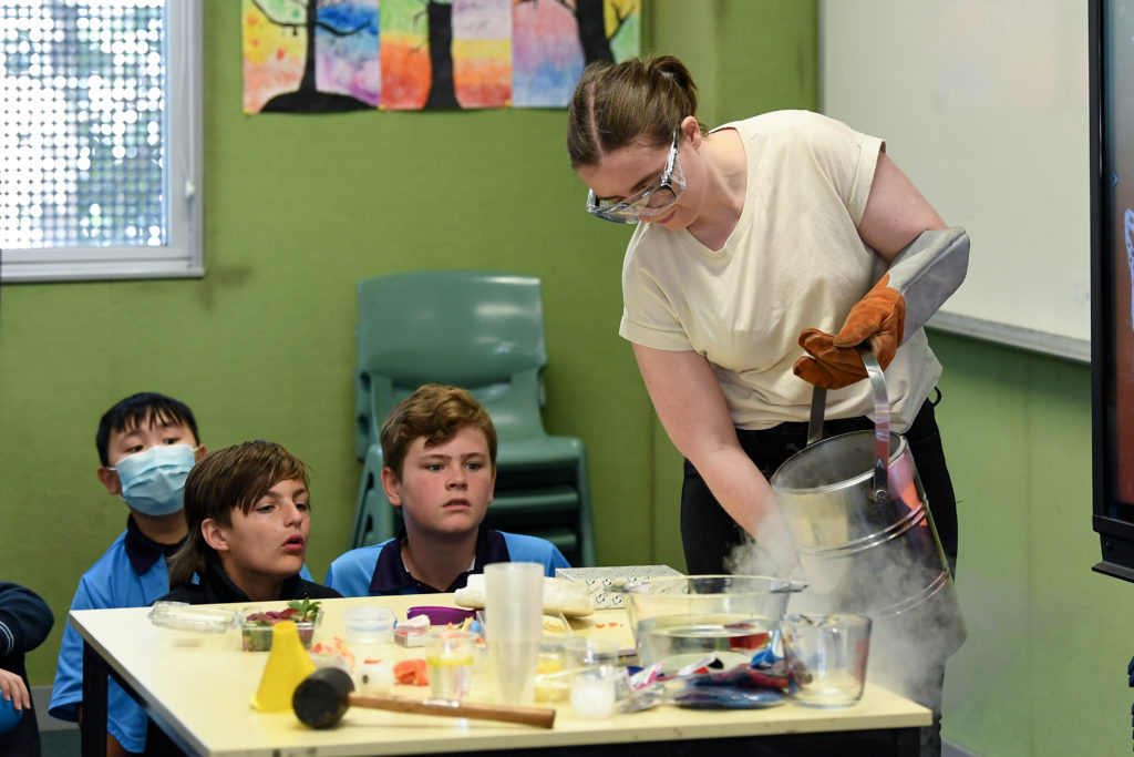 A woman in goggles runs a science experiment in a classroom as young children look on.