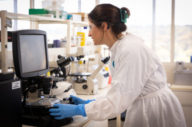 A woman in a lab coat and blue protective gloves examines data on a screen.