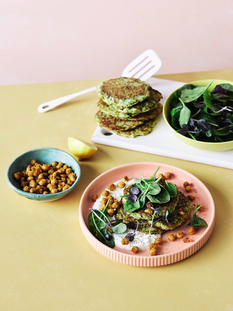 A stack of zucchini chickpea pancakes sit on a serving board next to a plate of mixed salad leaves. In front of the serving board is half a lemon, a bowl of crispy chickpeas and a plate with yoghurt dressing, zucchini pancakes, salad leaves and the crispy chickpeas scattered over the stack and plate.  