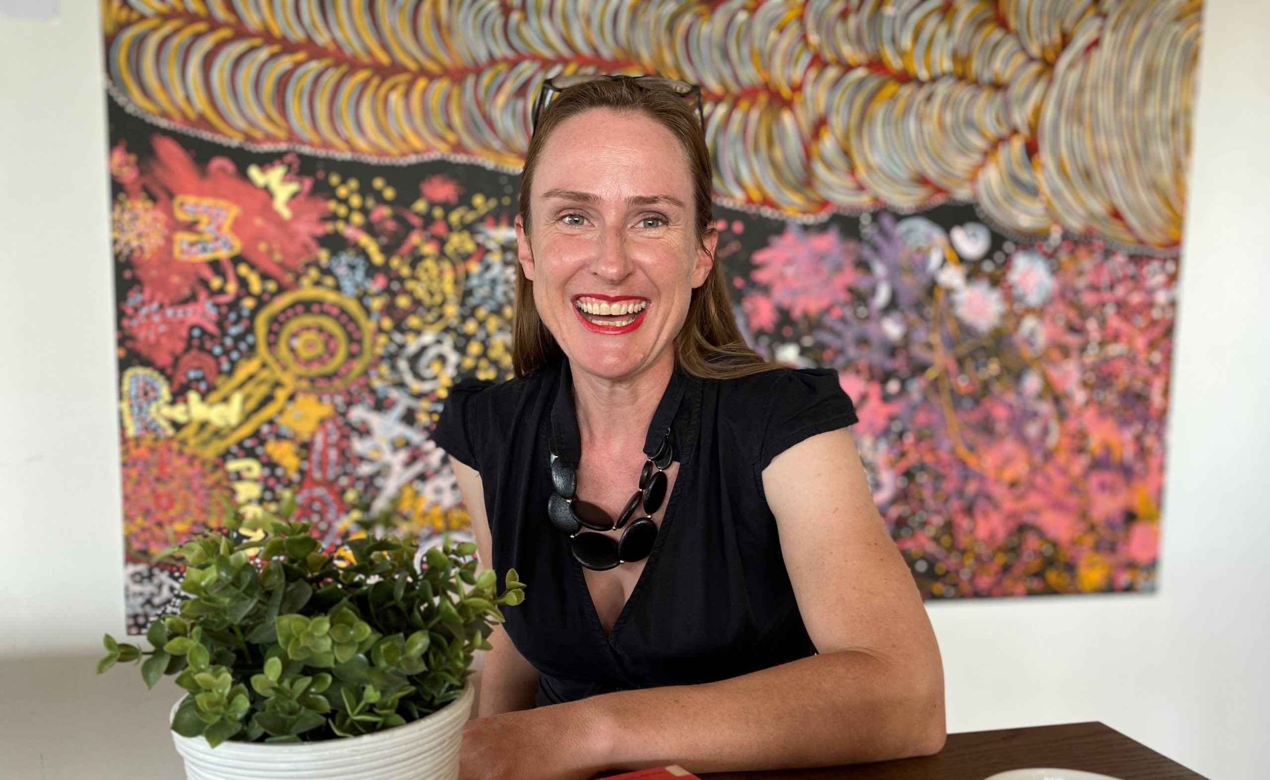 Portrait of Amelia Fyfield sitting in front of a colourful painting. She is smiling at the camera.