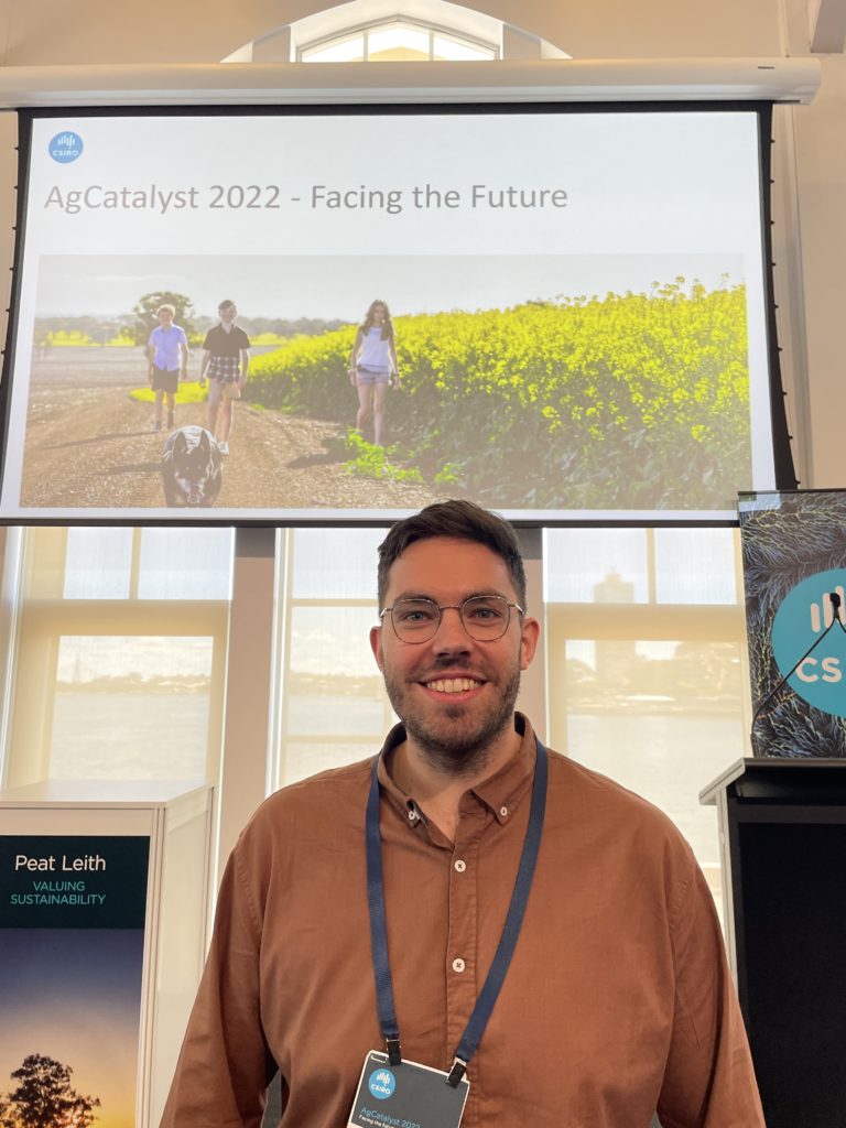 Person smiling at the camera, standing in front of a PowerPoint presentation slide that reads, AgCatalyst 2022 - Facing the Future. 