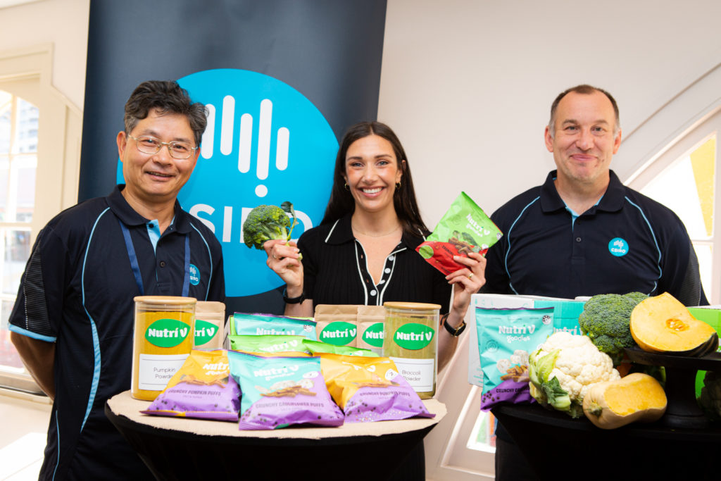 Three people standing behind a table with Nutri V products and vegetables. They're smiling at the camera. 