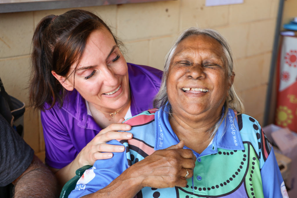 A woman in a purple shirt with long brown hair is smiling at and touching the shoulder of an older Wajarri woman wearing a blue shirt who is also smiling.