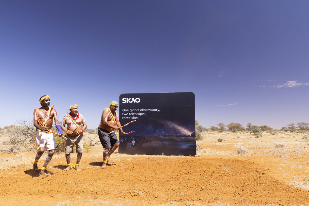 Three Indigenous men (Godfrey Simpson, Geoffrey Mongoo and Gerard Boddington) who are painted up are performing a Wajarri cultural dance in front of an SKAO sign. The sign reads: "SKAO One global observatory, two telescopes, three sites"