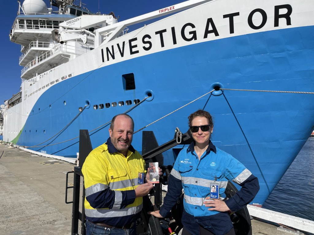 A male and female wearing hi-vis standing in front of the docked RV Investigator smiling, holding a coin