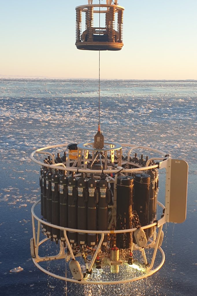 A cylindrical piece of scientific equipment covered in grey bottles hangs above the ocean from a wire.