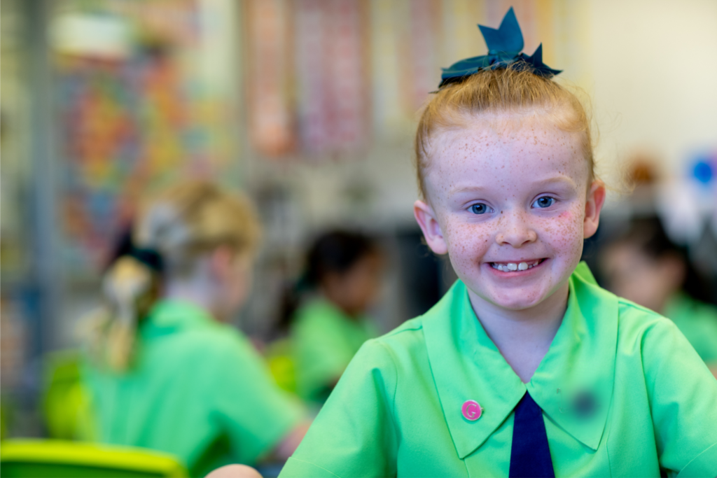 Girl in a school uniform smiling at the camera. Other students can be seen blurred in the background. 
