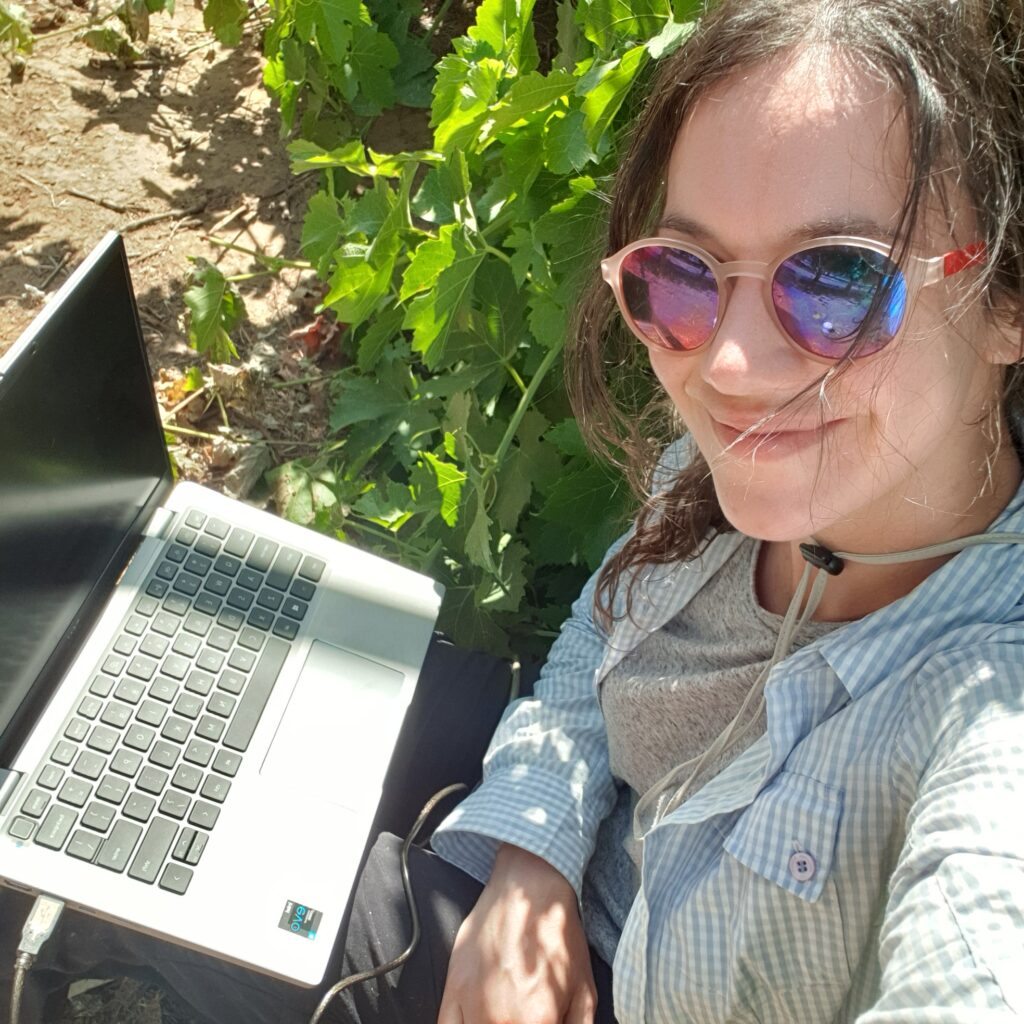 Selfie of a woman in sunglasses in a vineyard with a laptop on her knees.