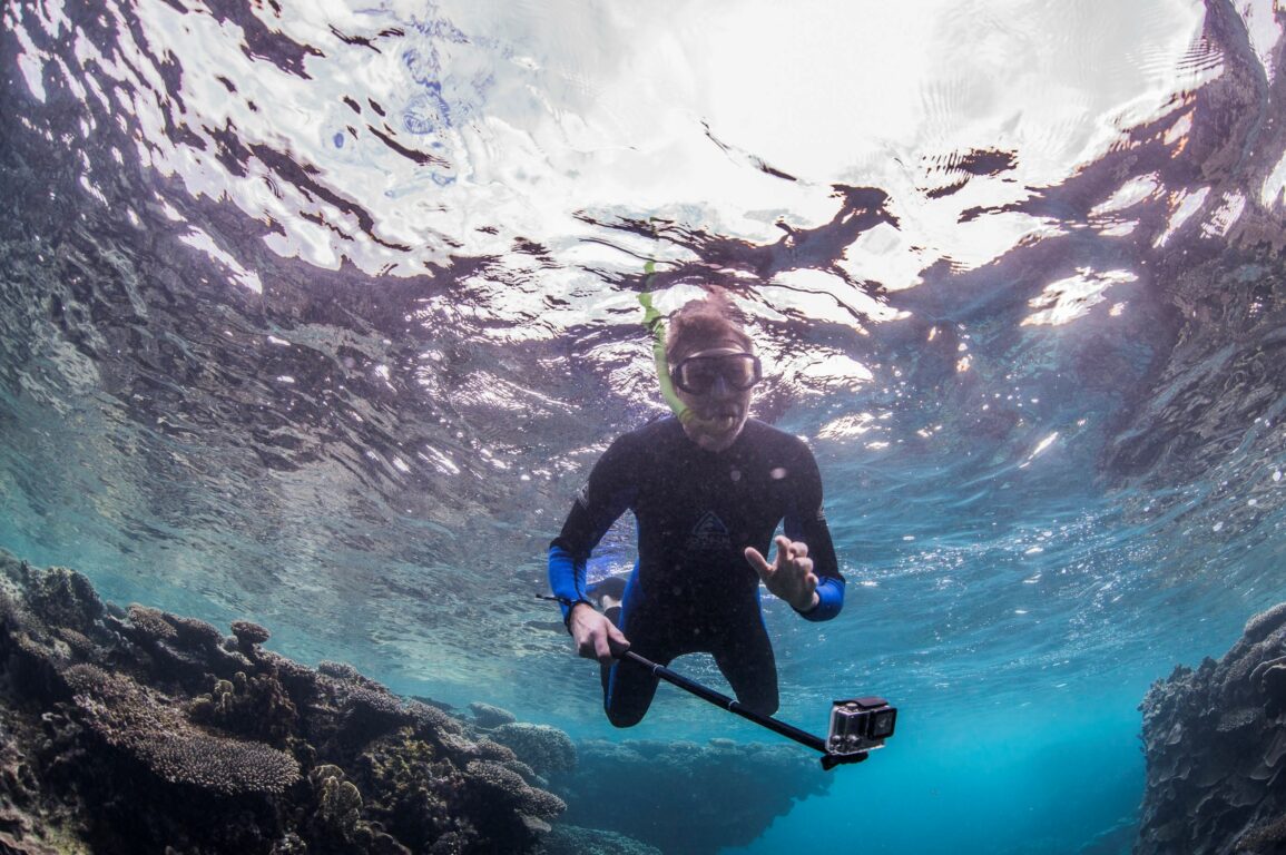 A man in a wetsuit snorkelling underwater above a reef holding a camera on a stick.
