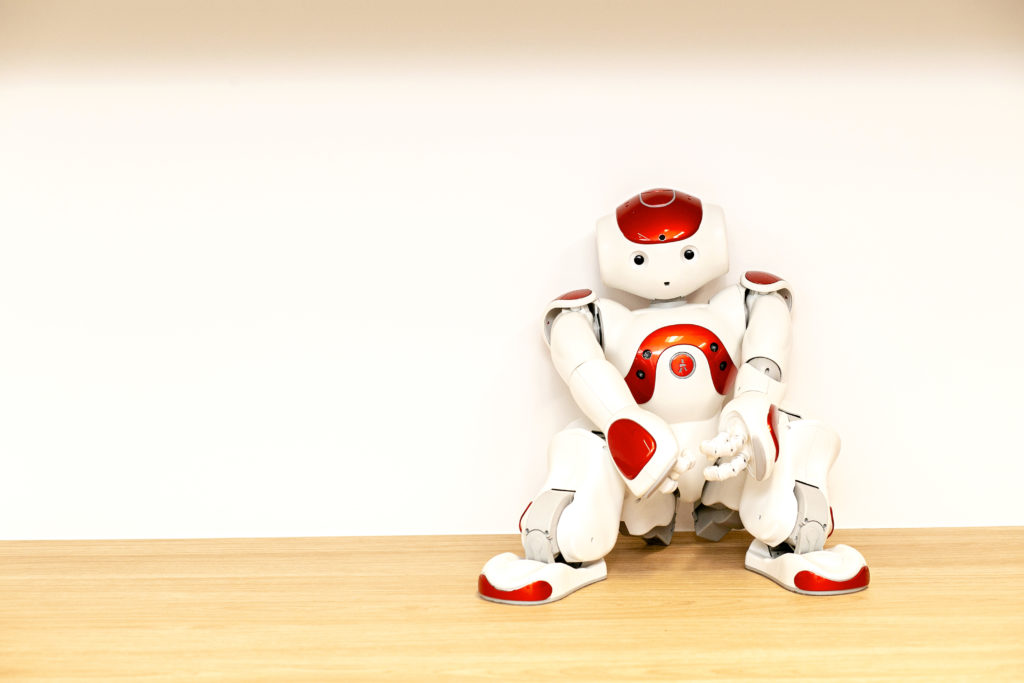 Photo of a small white and red robot sitting on the floor, leaning against a white wall.