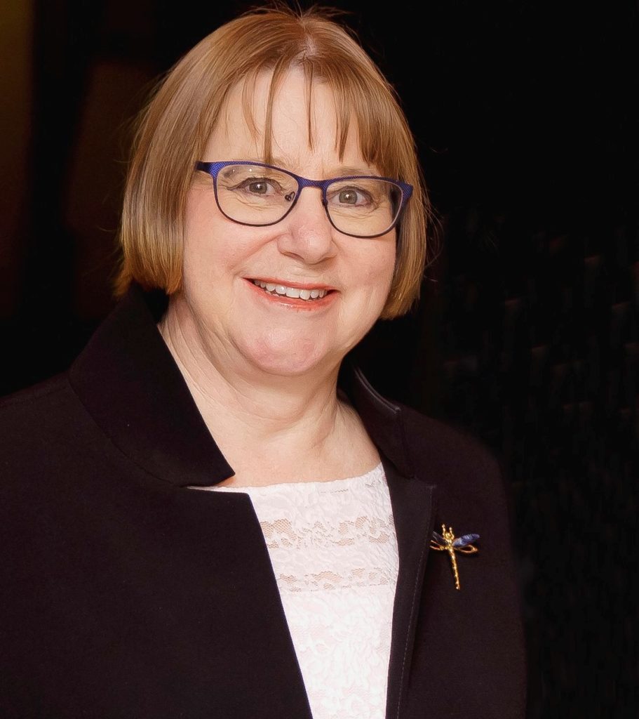 Portrait of smiling woman with short chestnut hair and blue-framed glasses wearing a white blouse and navy jacket wiith dragonfly pin. 