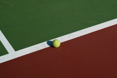 A yellow tennis ball on the white line of a green and clay-coloured tennis court