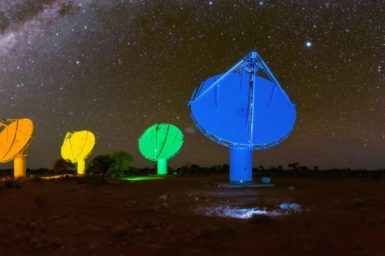 A photo taken at night showing a starry nights sky with five telescopes in the foreground. The telescopes are each illuminated in a different primary colour of the rainbow.