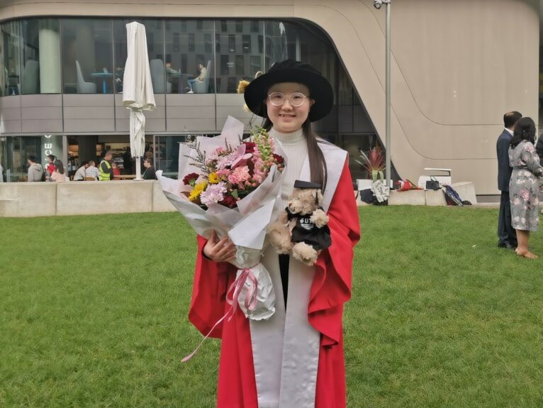 Lizhao poses in her graduation gown. She is carrying a large bouquet of flowers and a teddy bear.