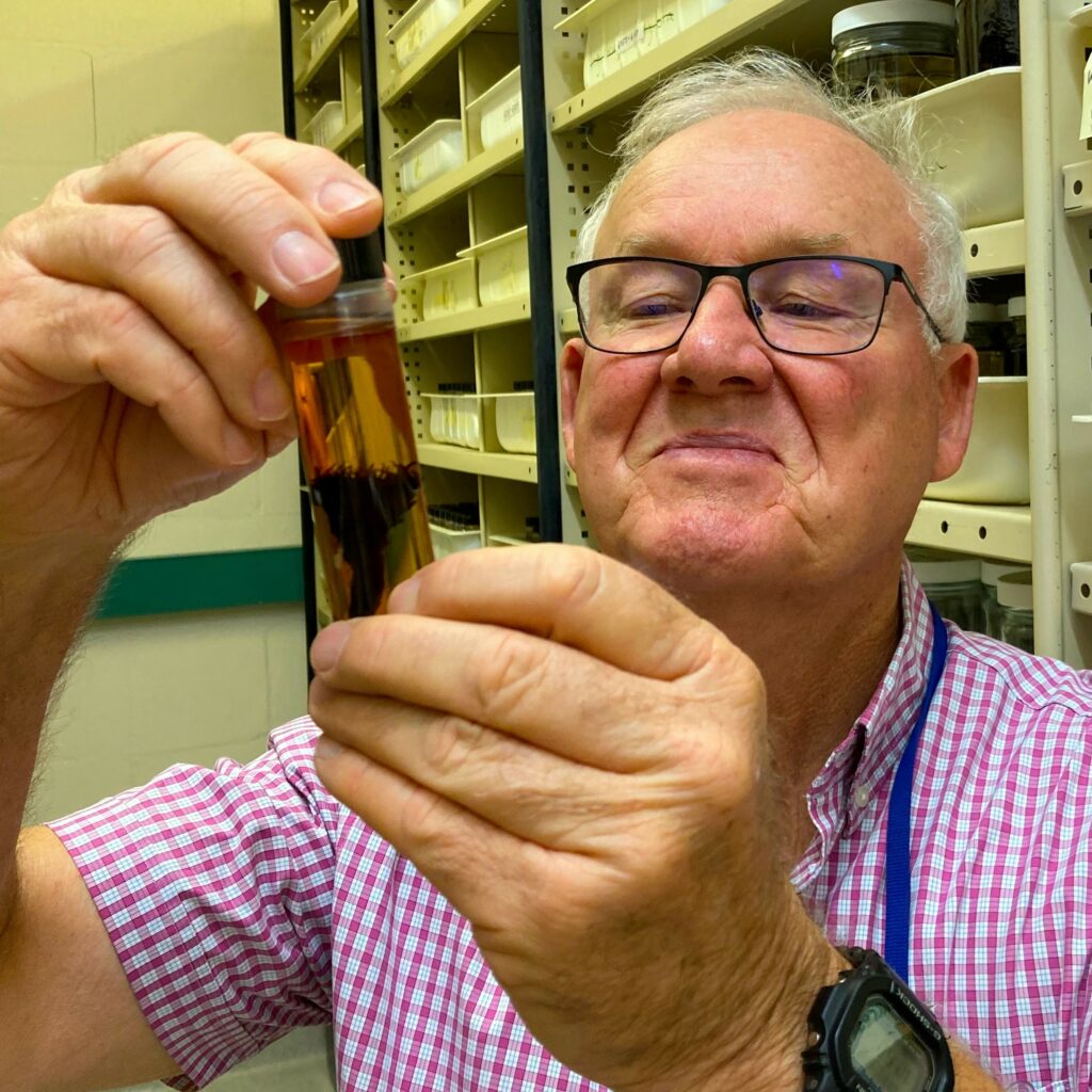 A man, Dr Mark Clements, holds up an orchid specimen preserved in ethanol in a tube. He is smiling at the orchid.