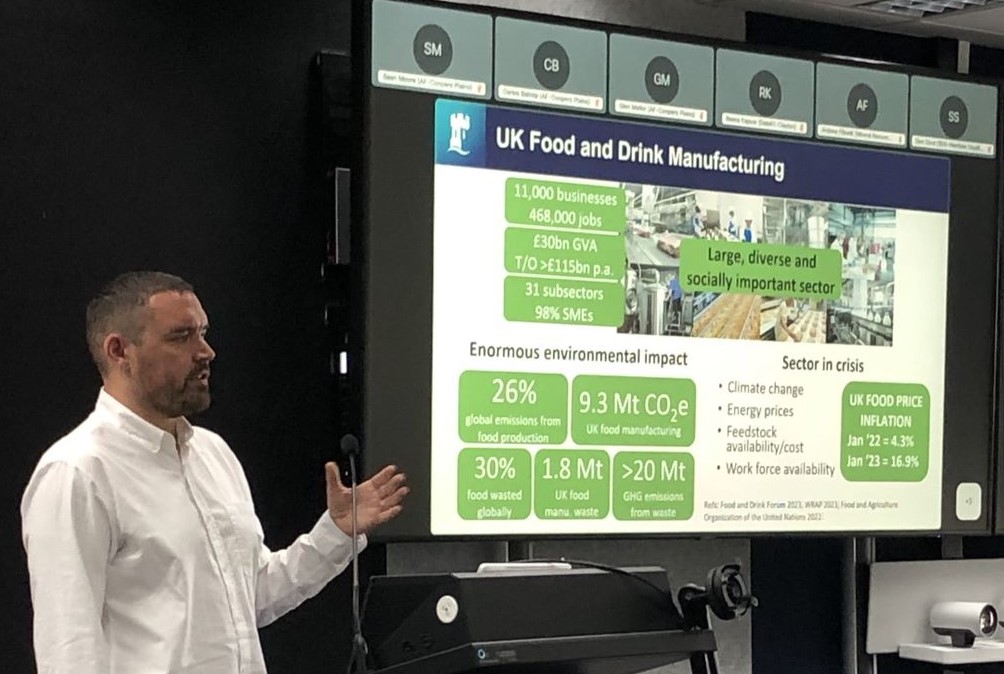 Assoc Prof Nik Watson from the University of Nottingham is standing at a microphone alongside a screen upon which is a slide titled UK Food and Drink Manufacturing.