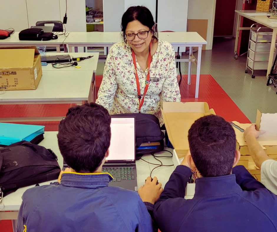 Veena, a middle aged woman leans over a desk while teaching two male students
